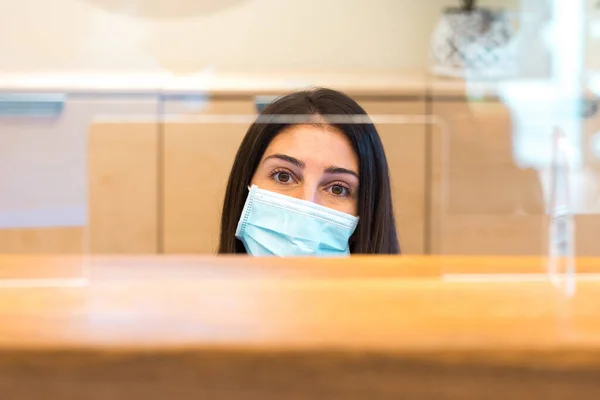 Donna che indossa una maschera facciale e seduta dietro la scrivania mentre lavora presso una reception di una spa o di un centro benessere. — Foto Stock