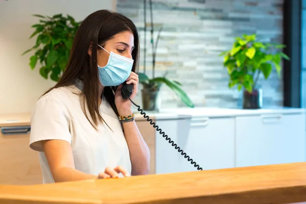 Il receptionist che indossa una maschera facciale riceve una chiamata mentre lavora presso una reception. — Foto Stock