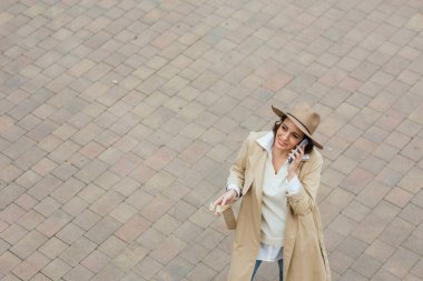 Şapkalı ve ceketli yetişkin bir kadın cep telefonuyla konuşuyor tuğla taş zeminde.