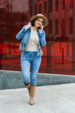 Yetişkin bir kadın, arkasında kırmızı camlar olan bir binaya karşı telefonla konuşuyor..