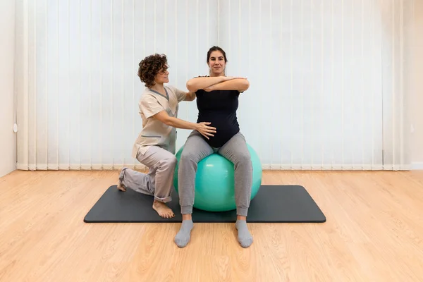 Egzersiz yapan hamile bir kadın, fizyoterapistinin yardımıyla bir fitness topunun üzerinde oturuyor. — Stok fotoğraf