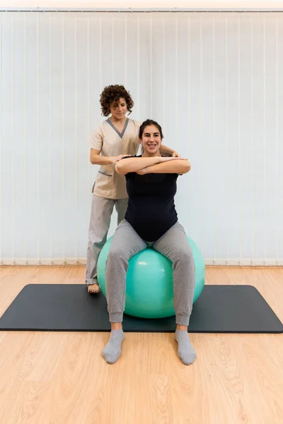 Una mujer embarazada haciendo ejercicios sentada en una pelota de fitness con los brazos delante con la ayuda de un fisioterapeuta —  Fotos de Stock