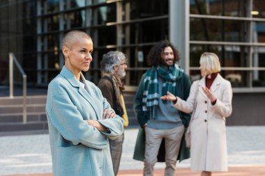Meslektaşları arka planda konuşurken dışarıda kollarını kavuşturmuş profesyonel bir iş kadını..