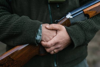 Kalın elbiseli bir adam tüfekle doğada duruyor.