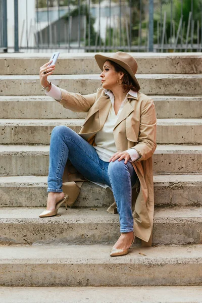 An elegant adult woman sitting on steps outdoors and taking a selfie — Stock Photo, Image