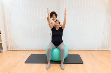 Hamile bir kadın fizyoterapistin yardımıyla bir fitness topunun üzerinde oturuyor.
