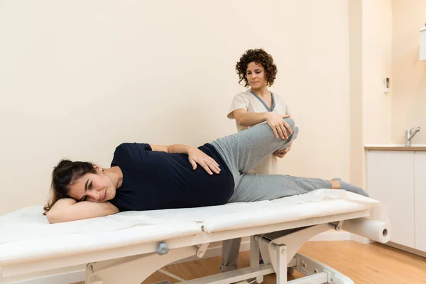 A pregnant woman lying on her side on a stretcher while a physiotherapist massages her legs at a health center. — Stock Photo, Image
