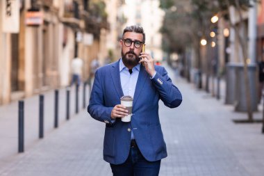 İş adamı elinde bir fincan kahve tutuyor ve sokakta yürürken telefonla konuşuyor..