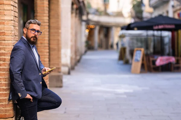 Yetişkin bir adam telefonuyla kameraya bakarak sütuna yaslanıyor.