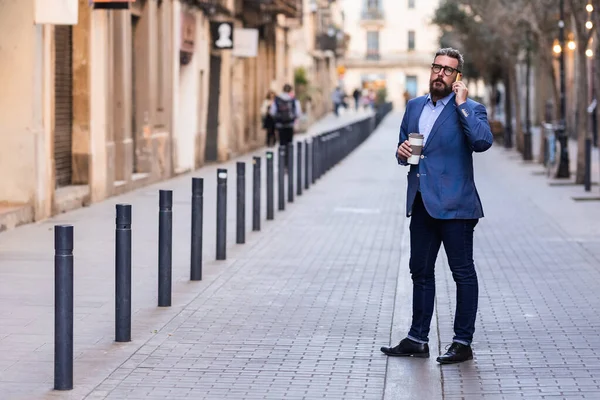 Хорошо одетый мужчина, стоящий в центре города и разговаривающий по телефону с кофейной чашкой — стоковое фото