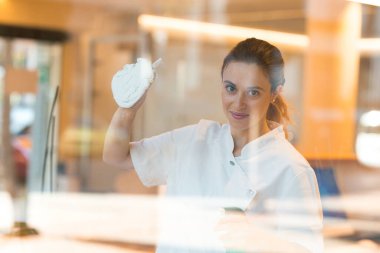 Temizlikçi kadın kameraya bakar ve bir sağlık merkezinin penceresini temizlerken gülümser..