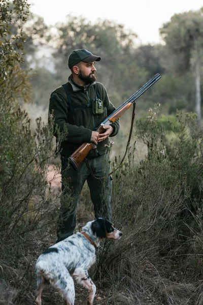 Hunter man with a gun standing outdoors with his dog while searching for a prey. — Stock Photo, Image