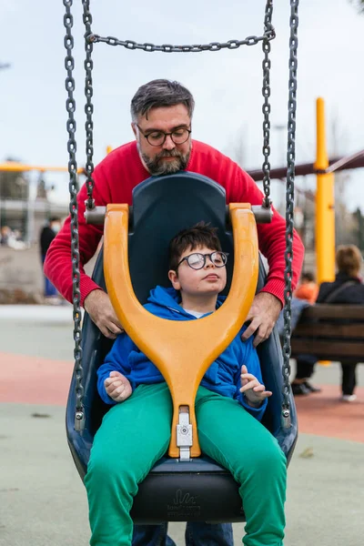 A relaxed child with a disability playing on adapted swing with his dad in a playground — 스톡 사진