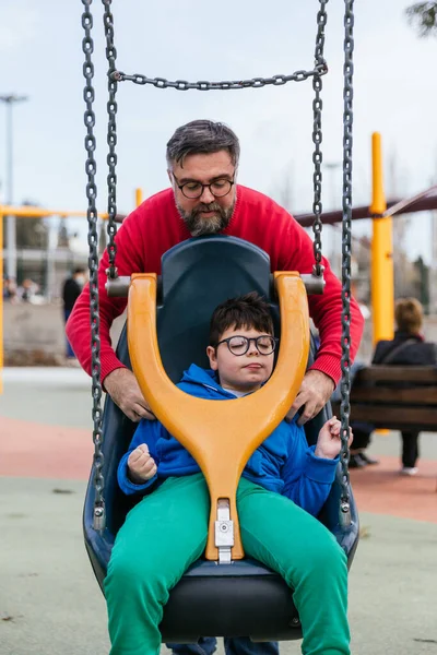Egy férfi, aki a mozgássérült fiát egy alkalmazkodott hintára kényszeríti a mozgássérültek számára a parkban lévő játszótéren.. — Stock Fotó