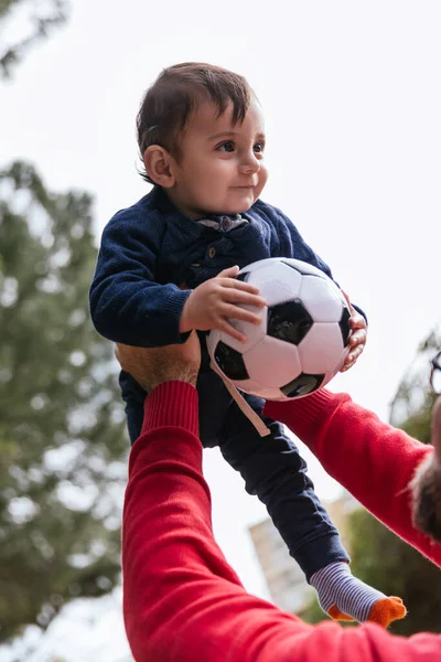 Father holding his little son up in the air with a soccer ball while playing together outdoors in the park. — 스톡 사진