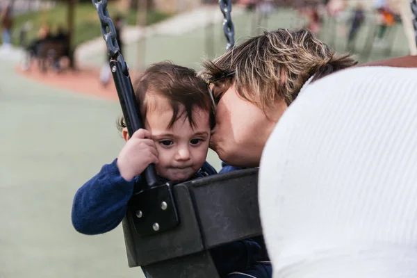 Lovely mom kissing her infant boy sitting on a swing at the playground — 스톡 사진