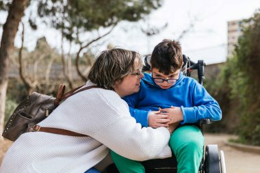 Parkta tekerlekli sandalyede engelli oğlunu kucaklayan sevgi dolu bir anne..
