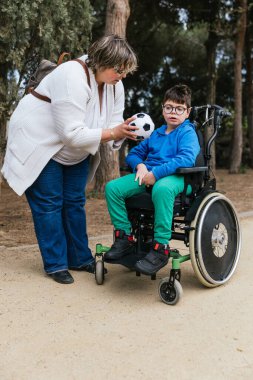 Parkta tekerlekli sandalyeyle oynayan engelli oğluna futbol topu veren bir anne.