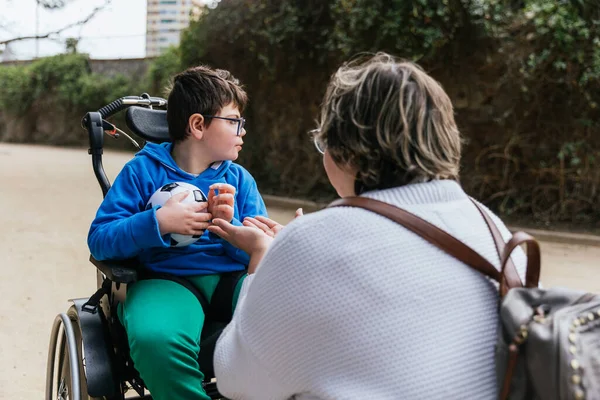 Tekerlekli sandalyede sakat bir çocuk elinde futbol topuyla parkta annesiyle oynuyor.