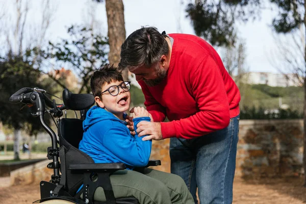 Parkta tekerlekli sandalye kullanan engelli oğluyla oynayan bir baba.