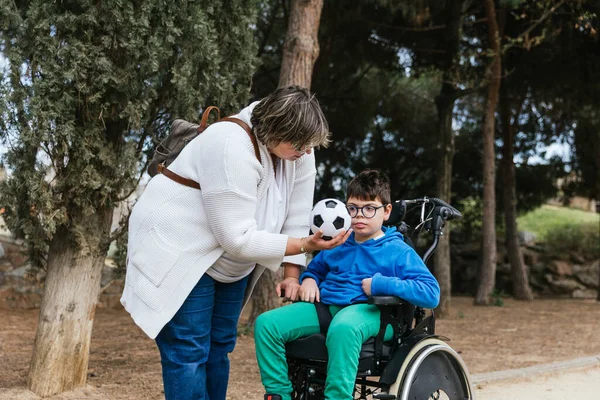 Egy anya focilabdát mutat a fogyatékos fiának, aki tolószéket használ a parkban.. Jogdíjmentes Stock Képek