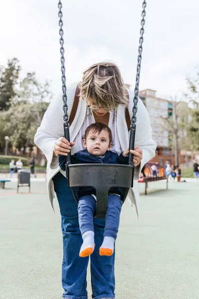 Mother pushing her little son in the swing in the playground in the park. — 스톡 사진