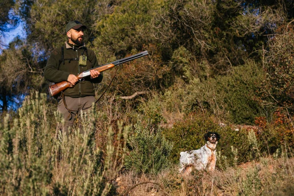 Jäger hält seine Waffe mitten in der Natur und schaut mit seinem Hund weg — Stockfoto