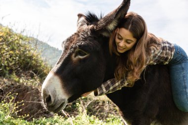 Mutlu genç bir kız eşeğe biniyor ve öne eğiliyor yüzünü okşuyor.
