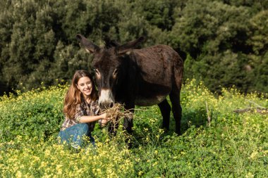 Genç bir kadın çayırda eşeği beslerken kameraya bakarak çömeliyor.
