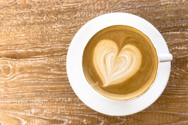 Ahşap masa arkasında kalp şeklinde sütlü sütlü sütlü sıcak kahve kapuçino bardağının en üst görüntüsü..