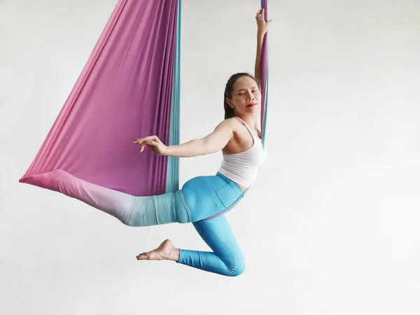 Mature woman pose in hammock performing aerial yoga or flying yoga exercise against white wall background in yoga studio room. Healthy lifestyles and emotional health of middle aged people concepts.