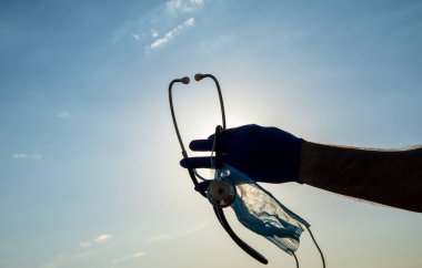 Mavi gökyüzüne karşı koruyucu bir maske ve steteskop tutan bir tıp doktoru. Coronavirus covid-19 enfeksiyonu.