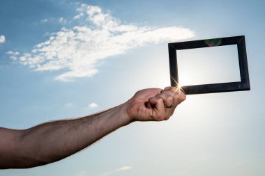 Mavi gökyüzünde bulutlu siyah bir resim ya da fotoğraf çerçevesi tutan eller. Bulanık plaj arka planında ilham verici bir fotoğraf çerçevesi.