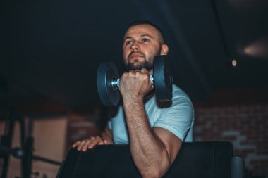 Formda ve kaslı bir adam koyu renk spor salonunda dambıllarla kol egzersizi yapıyor..
