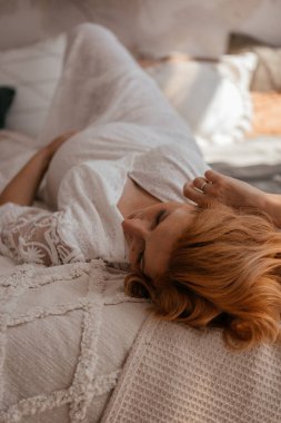 Avrupalı hamile kadın evdeki kanepede dinleniyor. Hamile kızıl saçlı kadın evde rahat bir yatakta uzanıyor..