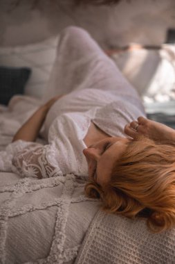 Avrupalı hamile kadın evdeki kanepede dinleniyor. Hamile kızıl saçlı kadın evde rahat bir yatakta uzanıyor..