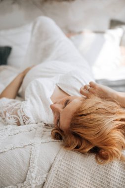 Avrupalı hamile kadın evdeki kanepede dinleniyor. Hamile kızıl saçlı kadın evde rahat bir yatakta uzanıyor..