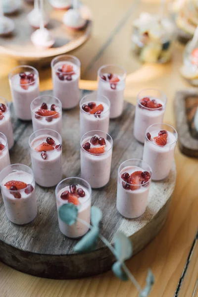 Mini cakes of fruit mousse decorated with strawberries and pomegranate seeds, standing on rustic wooden platter. Candy bar for a garden celebration. Natural and healthy sweets buffet. Selective focus.