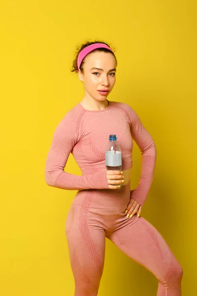 Hoş fitness kızı elinde şişeyle kameraya bakıyor, boş zaman, boş zaman, yaşam tarzı. İzole edilmiş sarı arka plan. Stüdyo. kopyalama alanı. Boş zaman
