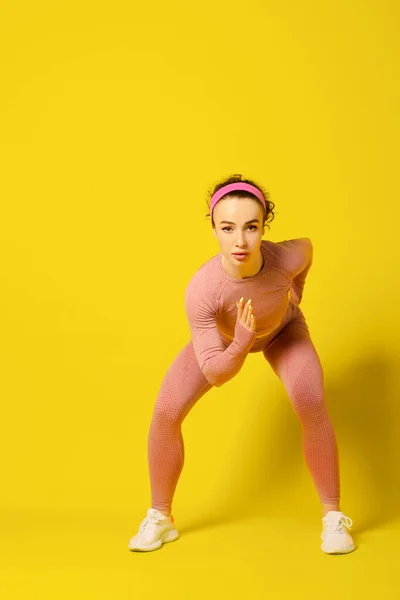 Antrenman yapıyorum. Pembe dar spor kıyafetleri içindeki genç kıvırcık kadın egzersiz yapıyor. Kopyala Space.Stüdyo resmi sarı arka planda izole edildi.