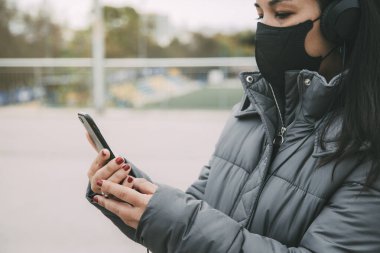 Koruyucu maskeli, kulaklık takan, cep telefonu kullanan Asyalı genç bir kadın. Boşluğu kopyala.