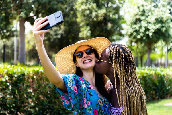 Unga multiraciala par homosexuella kvinnor tar selfie och kysser på kinden i parken. — Stockfoto