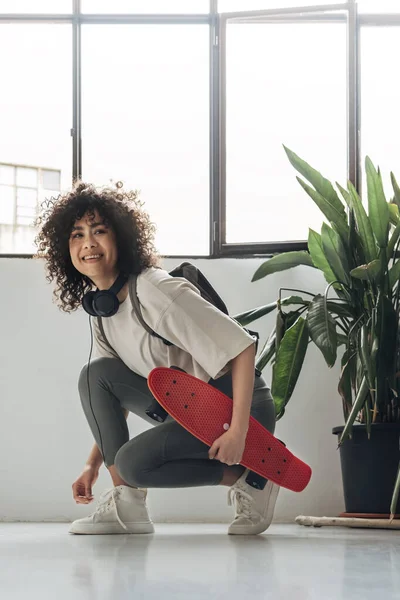 Leende ung multiracial kvinna binda sneaker skosnöre medan du håller skateboard och bär hörlurar. Vertikal bild. — Stockfoto