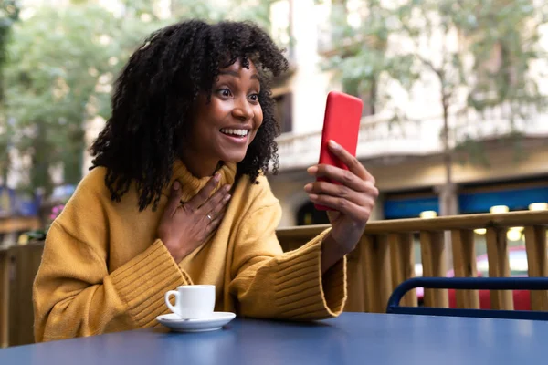 Joven mujer afroamericana con expresión sorpresa en videollamada usando smartphone en cafetería al aire libre.Espacio para copiar. —  Fotos de Stock