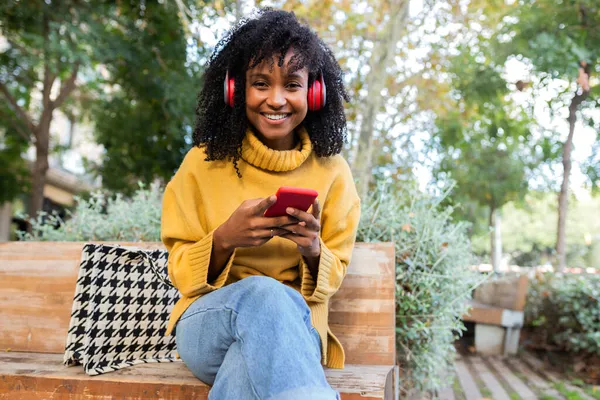 Ung afroamerikansk kvinna i hörlurar med mobiltelefon sitter på parkbänken och tittar på kameran. Kopiera utrymme. — Stockfoto