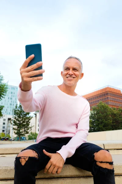 Vit vuxen man med rosa tröja som tar en selfie utomhus med hjälp av smartphone. Vertikal bild. — Stockfoto