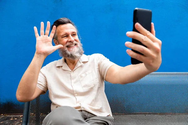 Vit mogen man med skägg som sitter på en bänk och vinkar hej på videosamtal. Blå bakgrund. — Stockfoto