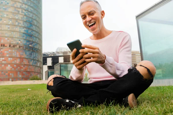Le kaukasisk man sitter på gräs i stadens offentliga park tittar på mobiltelefon. — Stockfoto