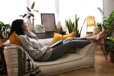 Beyaz bir kadın kanepede uzanıp evde kitap okuyor. Boşluğu kopyala.