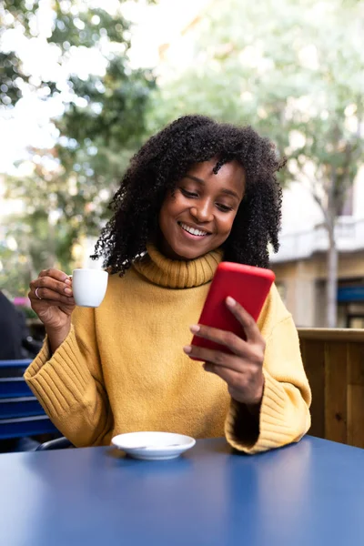 Glad ung afroamerikansk kvinna som använder mobiltelefon dricker kaffe i en utomhus kafé terrass. Vertikal bild. — Stockfoto
